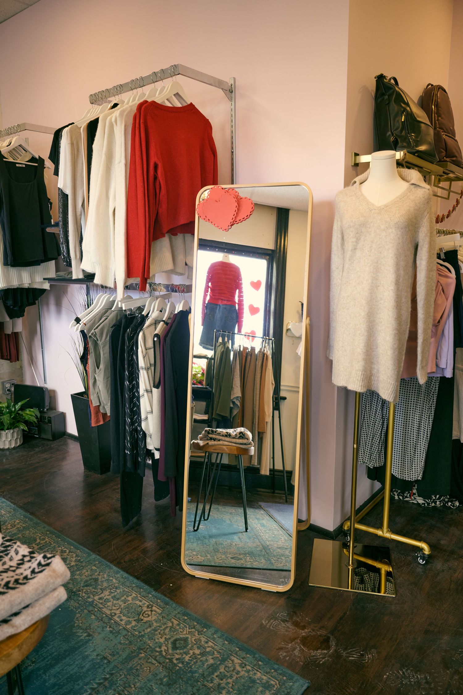 Interior of a boutique clothing store showing hanging garments, a full-length mirror, and wooden floors with a decorative rug.