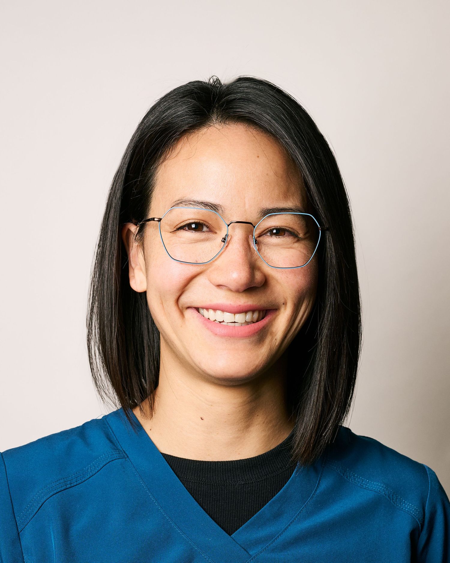 Smiling woman with glasses and dark hair wearing teal medical scrubs against a light background.