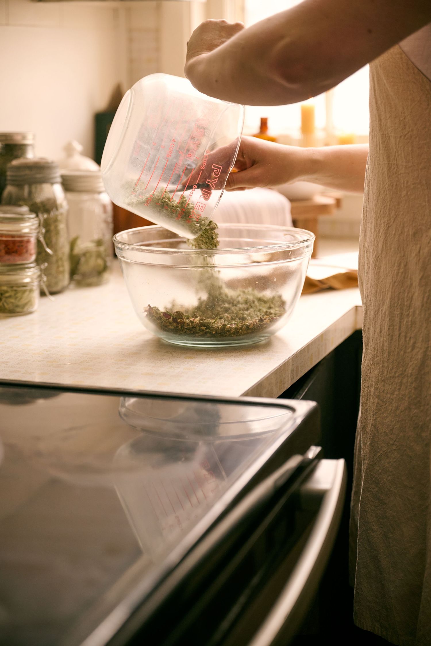 Hands pouring dried herbs from a container into a glass mixing bowl on a kitchen counter with mason jars in the background.