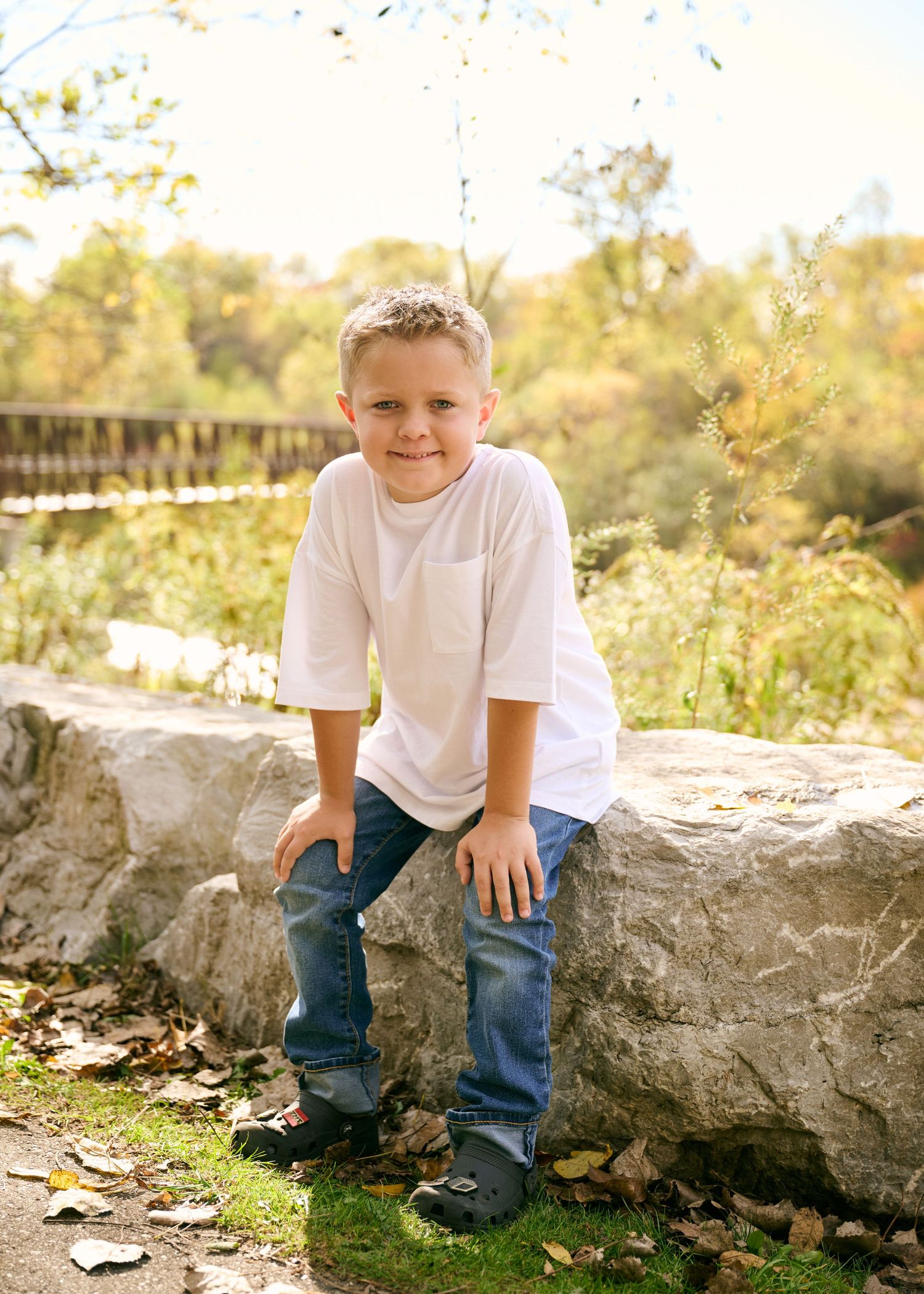A young person in a white t-shirt and jeans sits on a large rock in a sunny outdoor setting with autumn foliage.