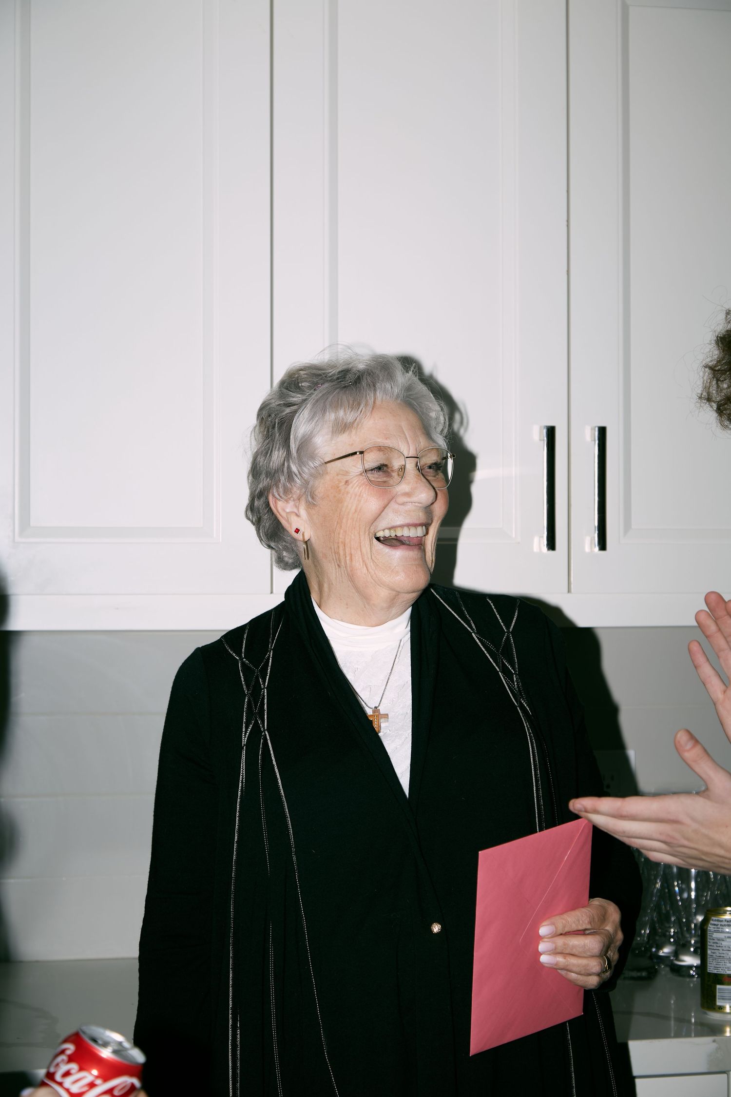 Elderly person wearing glasses and a black cardigan laughs joyfully while holding a red folder in a kitchen setting.