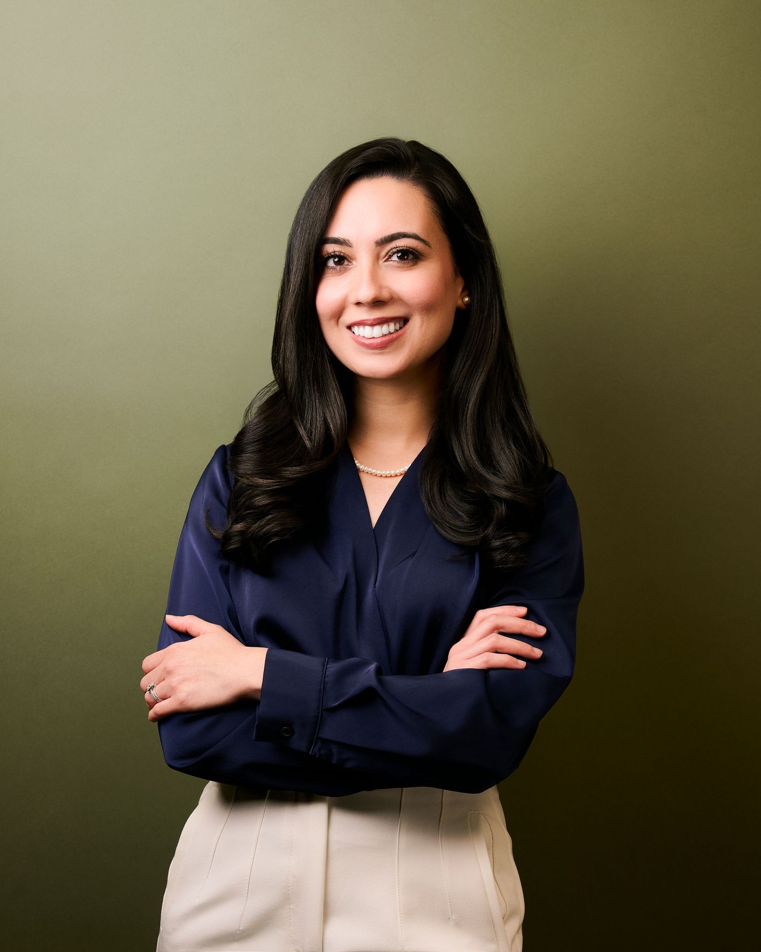 Professional woman in navy blouse with arms crossed, smiling confidently against olive green background.