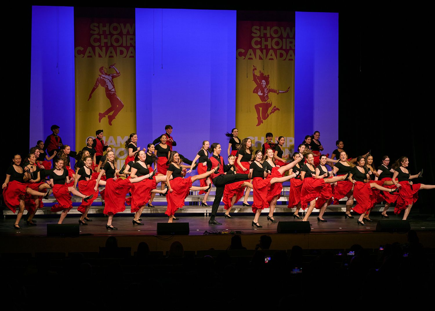 Performers in red and black costumes dance on stage at Show Choir Canada competition under bright spotlights.