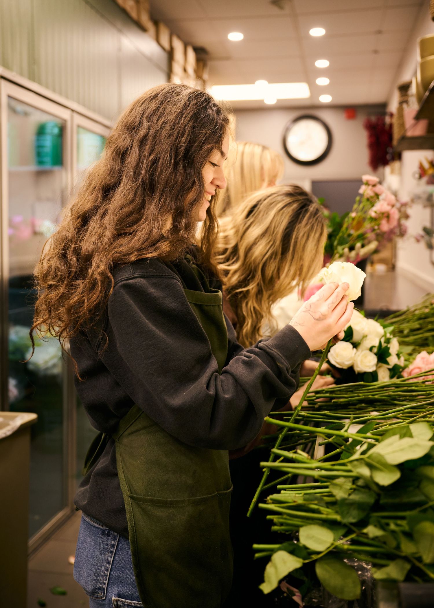 Florist in a green apron examines fresh white roses against a backdrop of flowers and greenery in a shop interior.