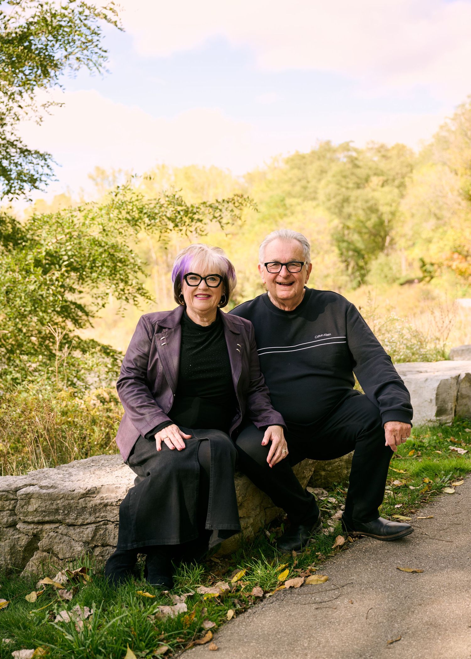 Two people sit together on a stone wall along a path surrounded by autumn foliage on a sunny afternoon.