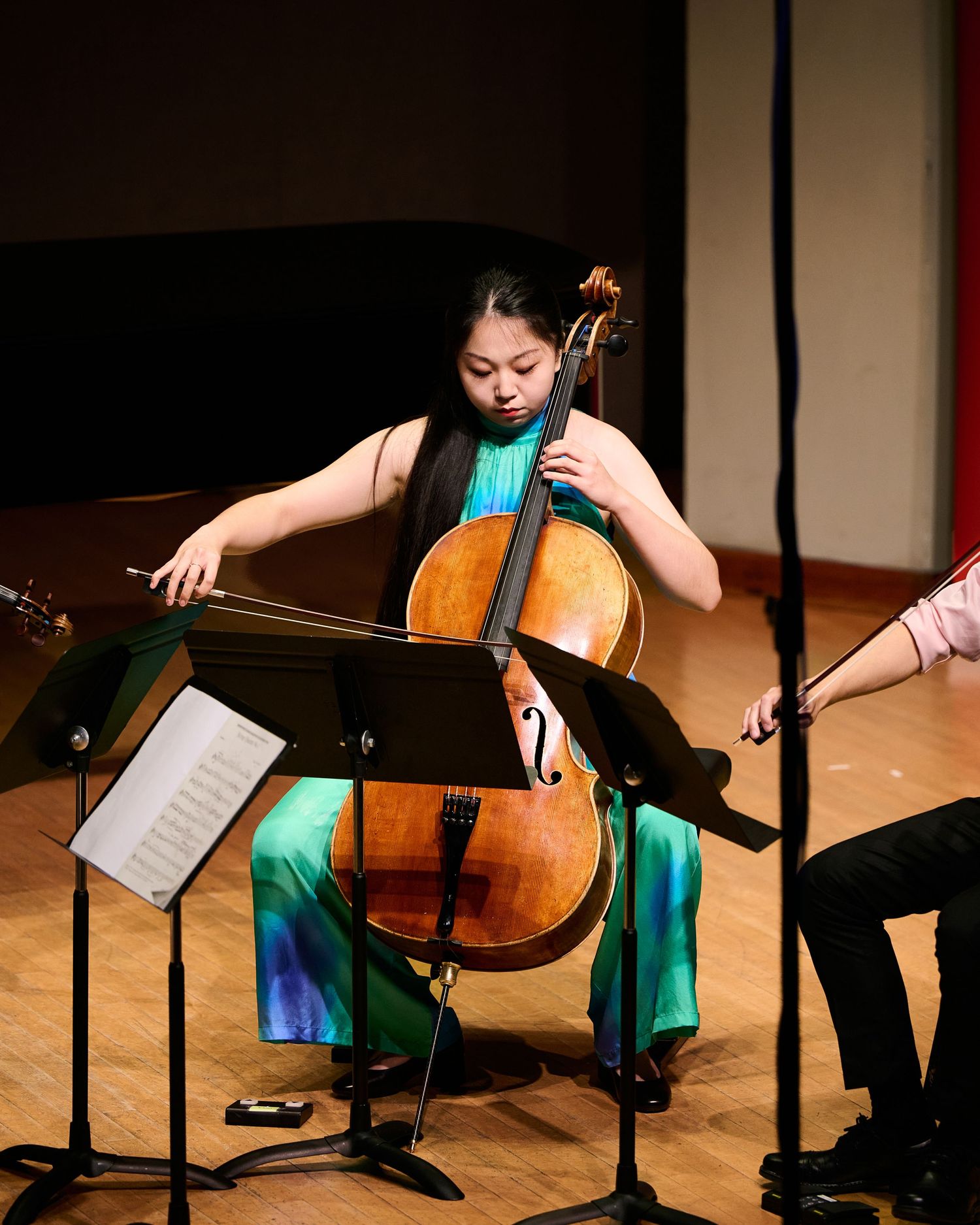 Young female cellist performing on stage in a teal outfit, playing cello with sheet music on a stand.