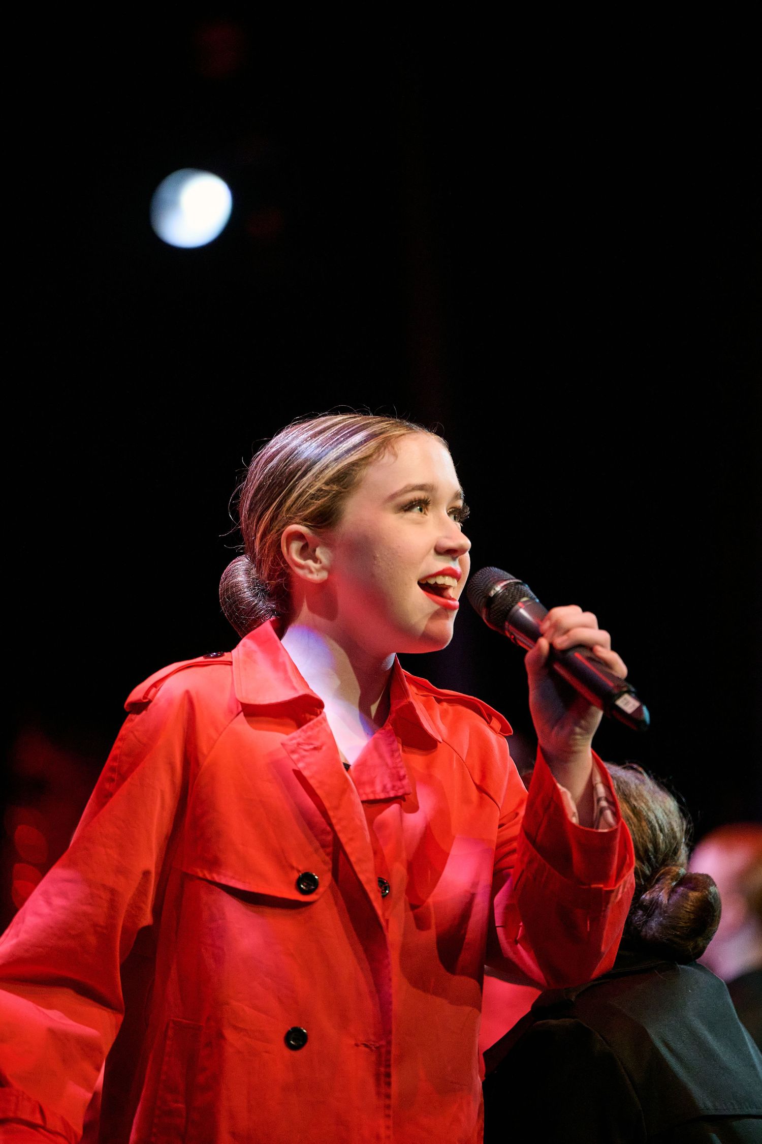 A performer singing on stage wearing a bright red coat under dramatic spotlight illumination.