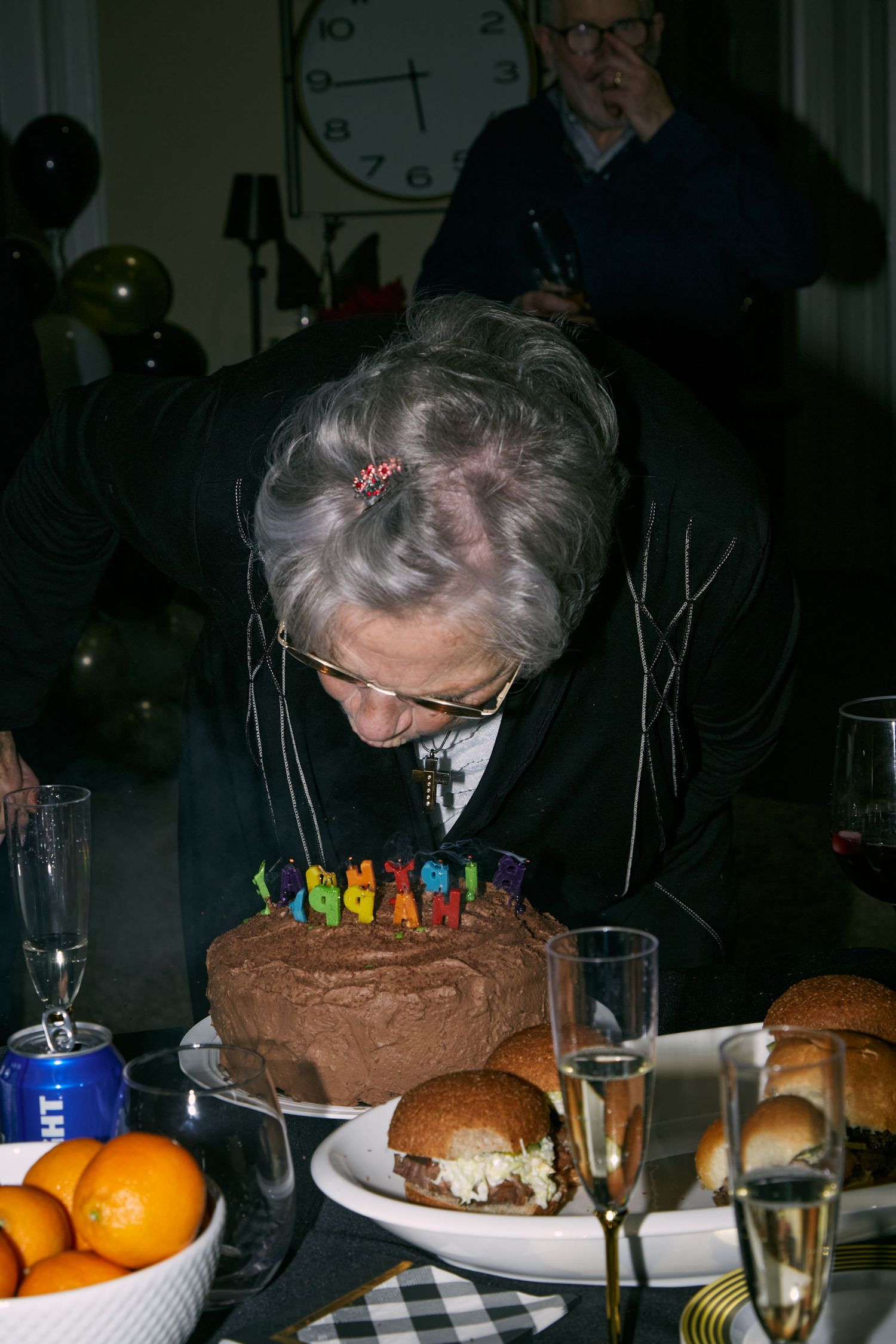 Someone blowing out birthday candles on a chocolate cake at a dinner celebration with oranges and other food on the table.