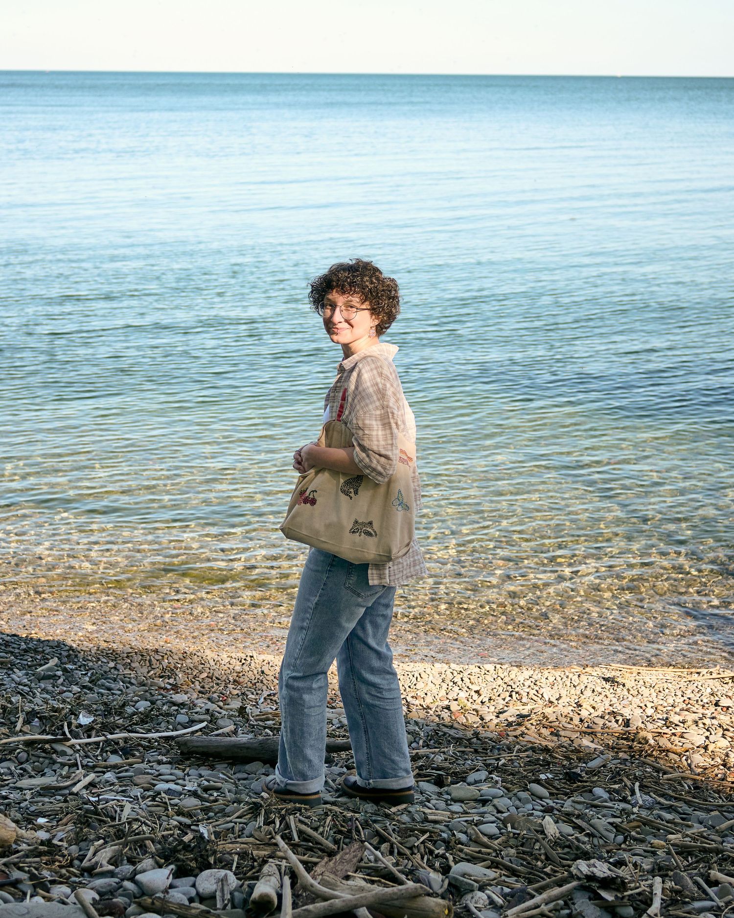 Someone stands on a rocky beach shoreline wearing casual clothes and a shoulder bag with the blue ocean waters in the background.