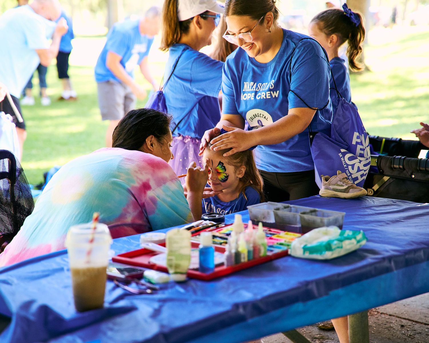Volunteers in blue shirts paint a child's face at an outdoor community event with art supplies on a blue table.