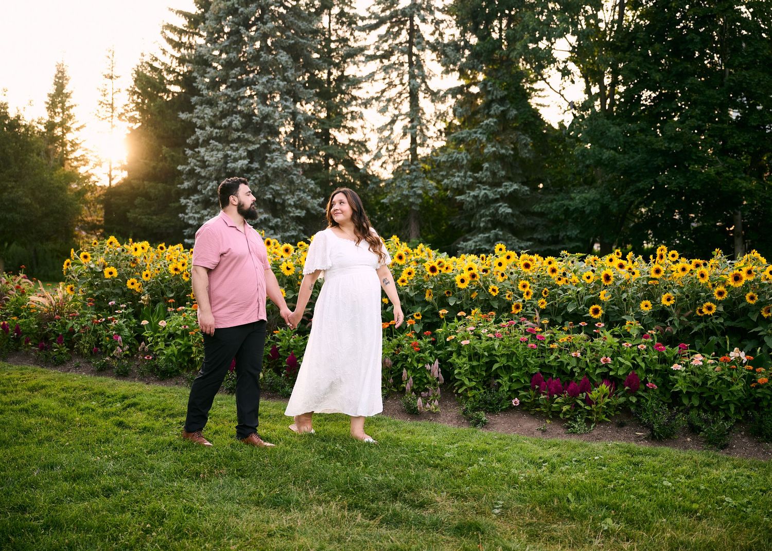 Couple holding hands in front of vibrant sunflower garden at golden hour sunset in lush green park.
