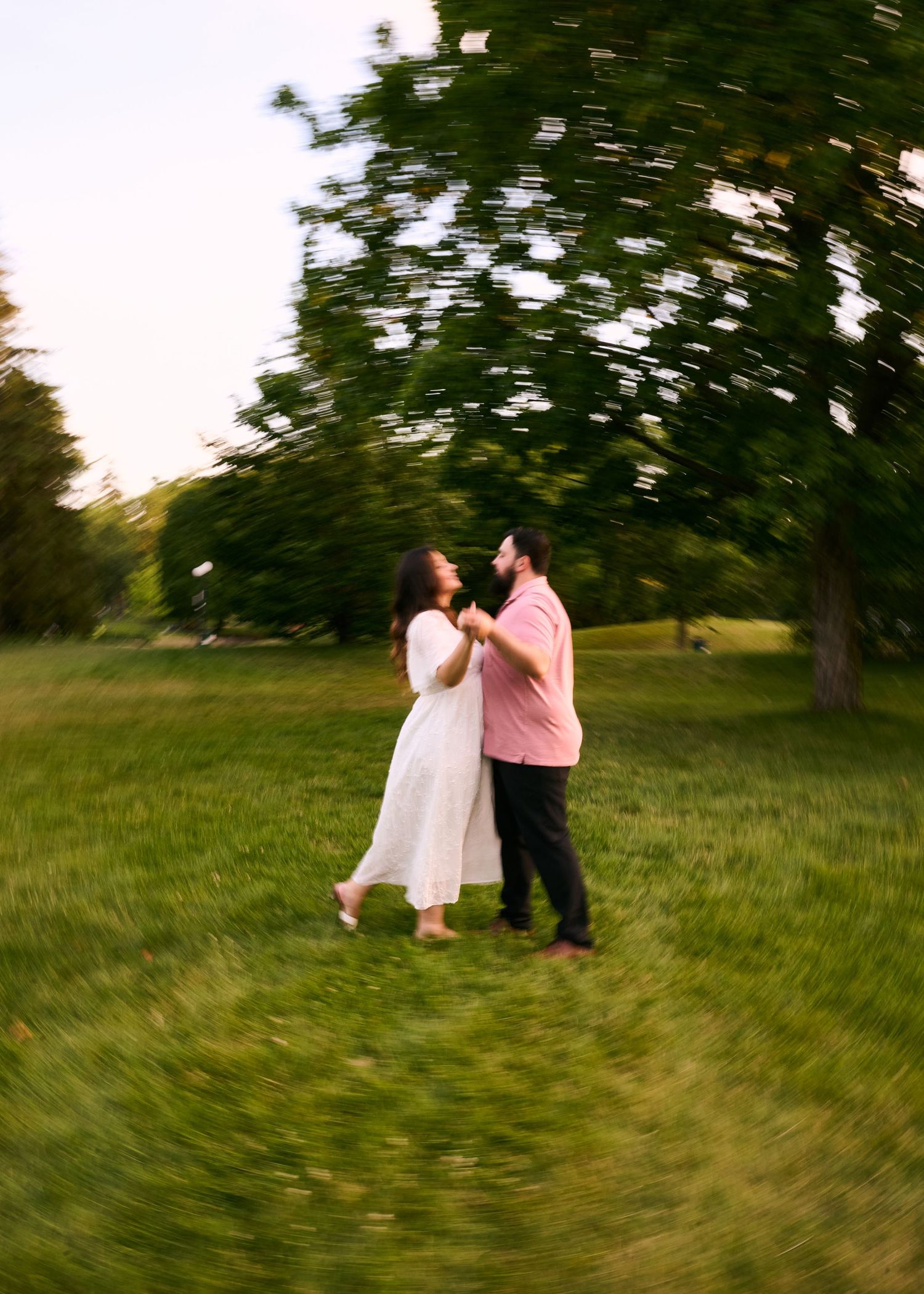 Couple dancing together in a lush green park at sunset, woman in white dress and man in pink shirt.