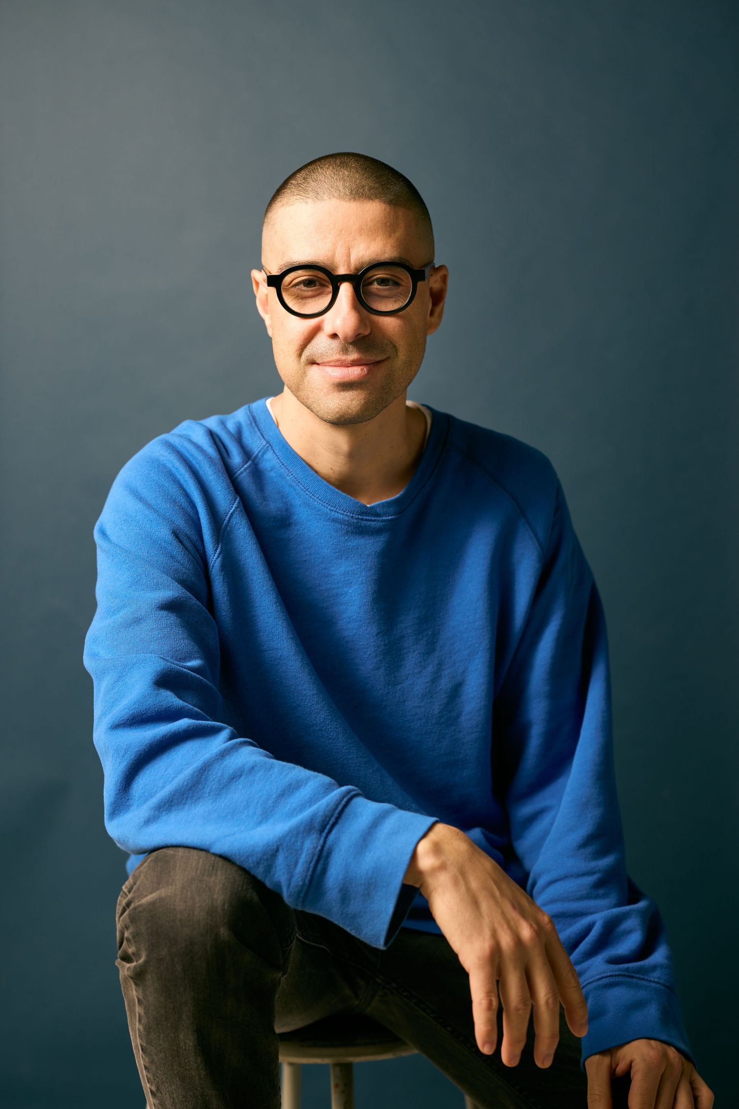 A person wearing glasses and a bright blue sweater sits casually on a chair against a dark background.