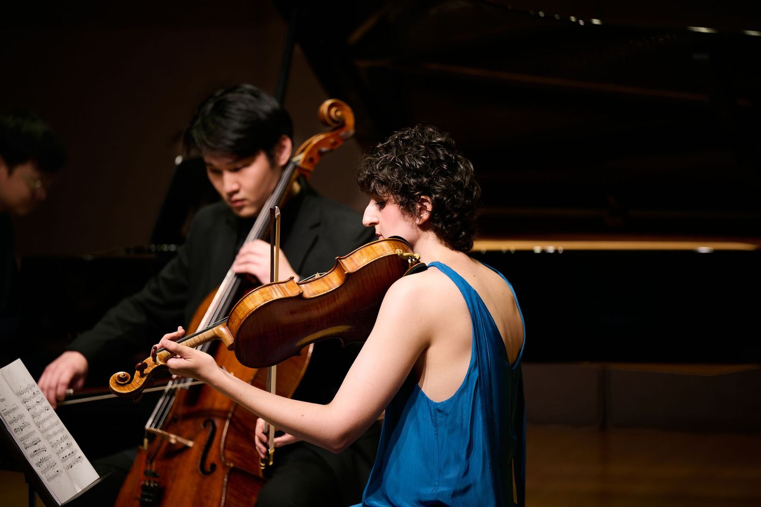 Two musicians perform chamber music, a cellist and violinist in elegant attire on a concert stage with a grand piano.
