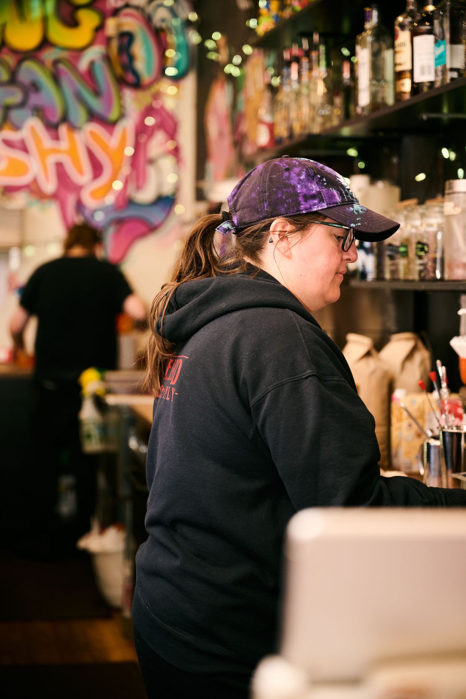 A bar worker prepares drinks against a vibrant graffiti wall with string lights in the background.