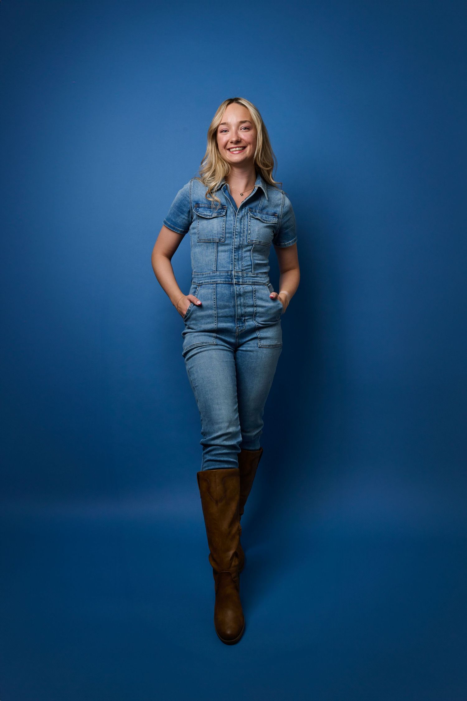 Young woman in denim jumpsuit and brown boots poses confidently against blue backdrop.