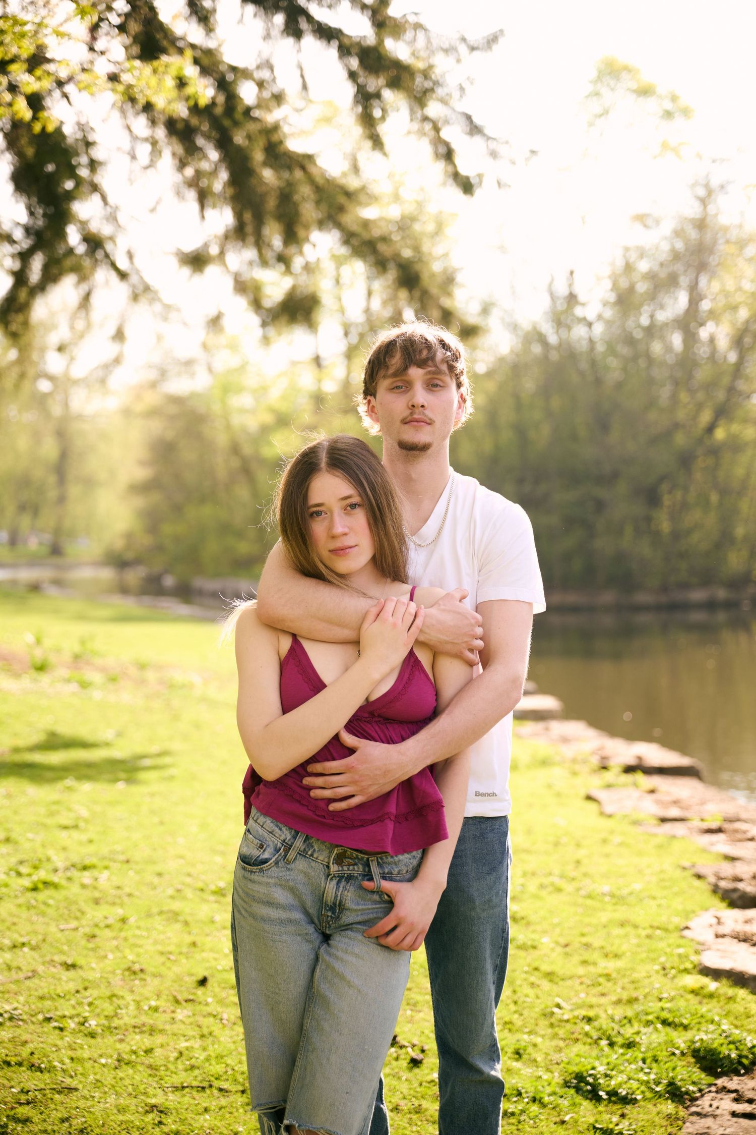Young couple embracing in a sunny park by a pond, woman in purple top and jeans, man in white t-shirt.