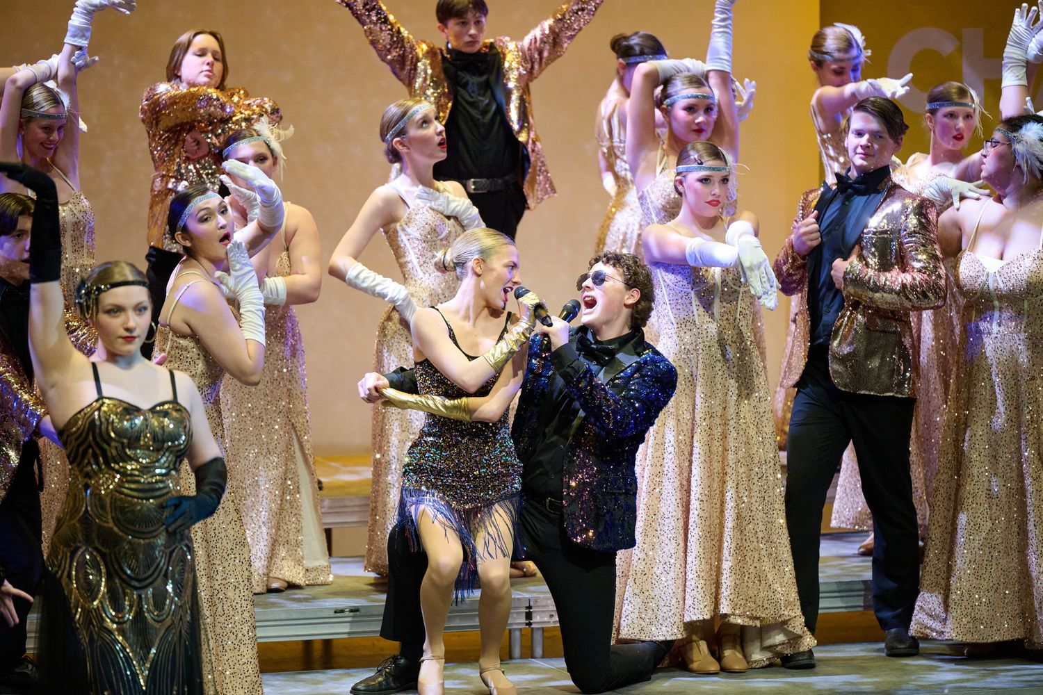 Performers singing and dancing together on stage in sparkly gold dresses and formal attire during a theatrical production.