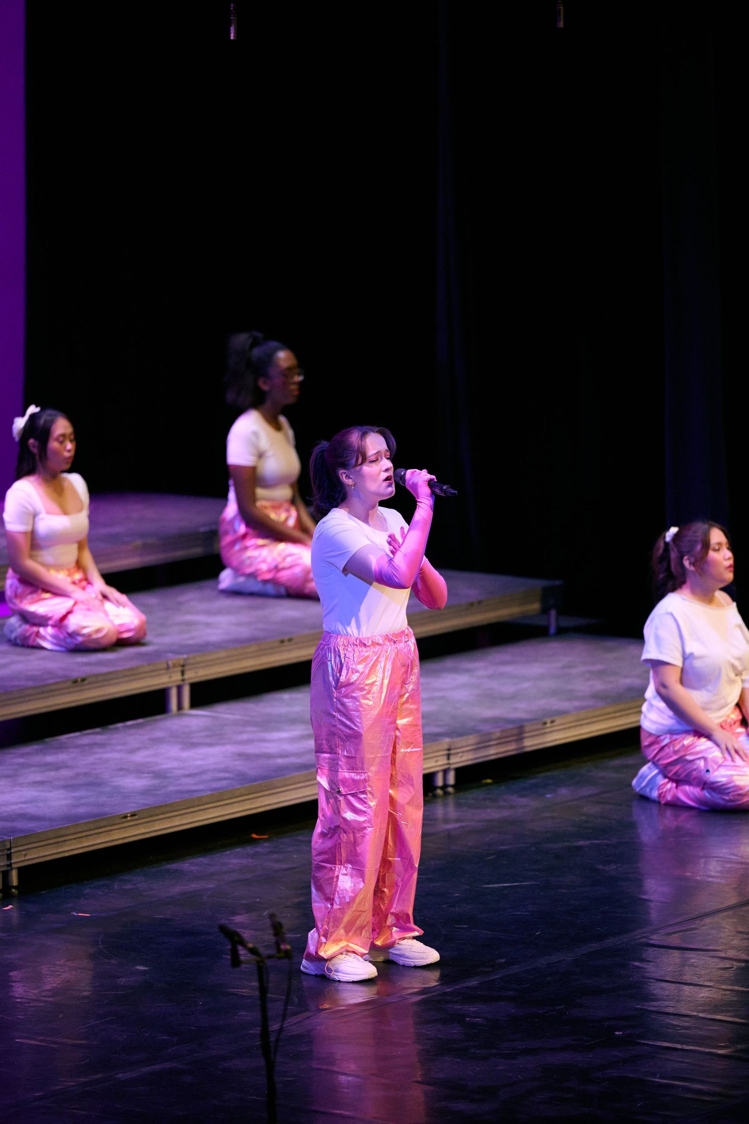 Performers sit and kneel on wooden platforms during a stage production with dramatic purple lighting.
