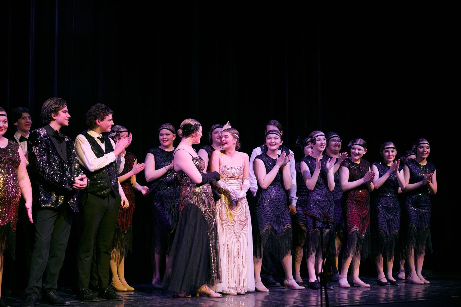 Performers in dark attire stand in a line formation on stage during a theatrical production.