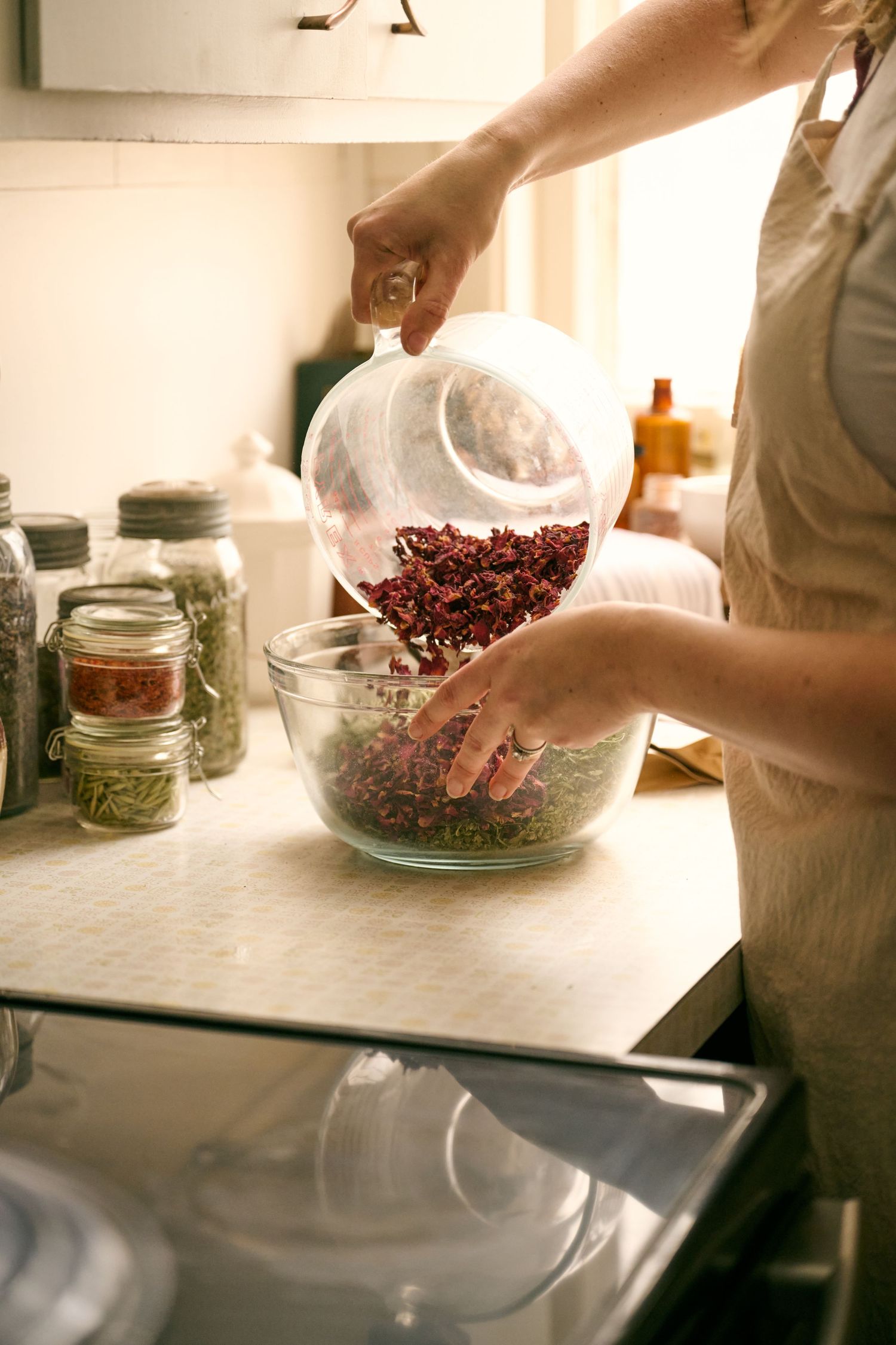 Hands pouring dried cranberries from a white container into a glass mixing bowl in a sunlit kitchen setup.