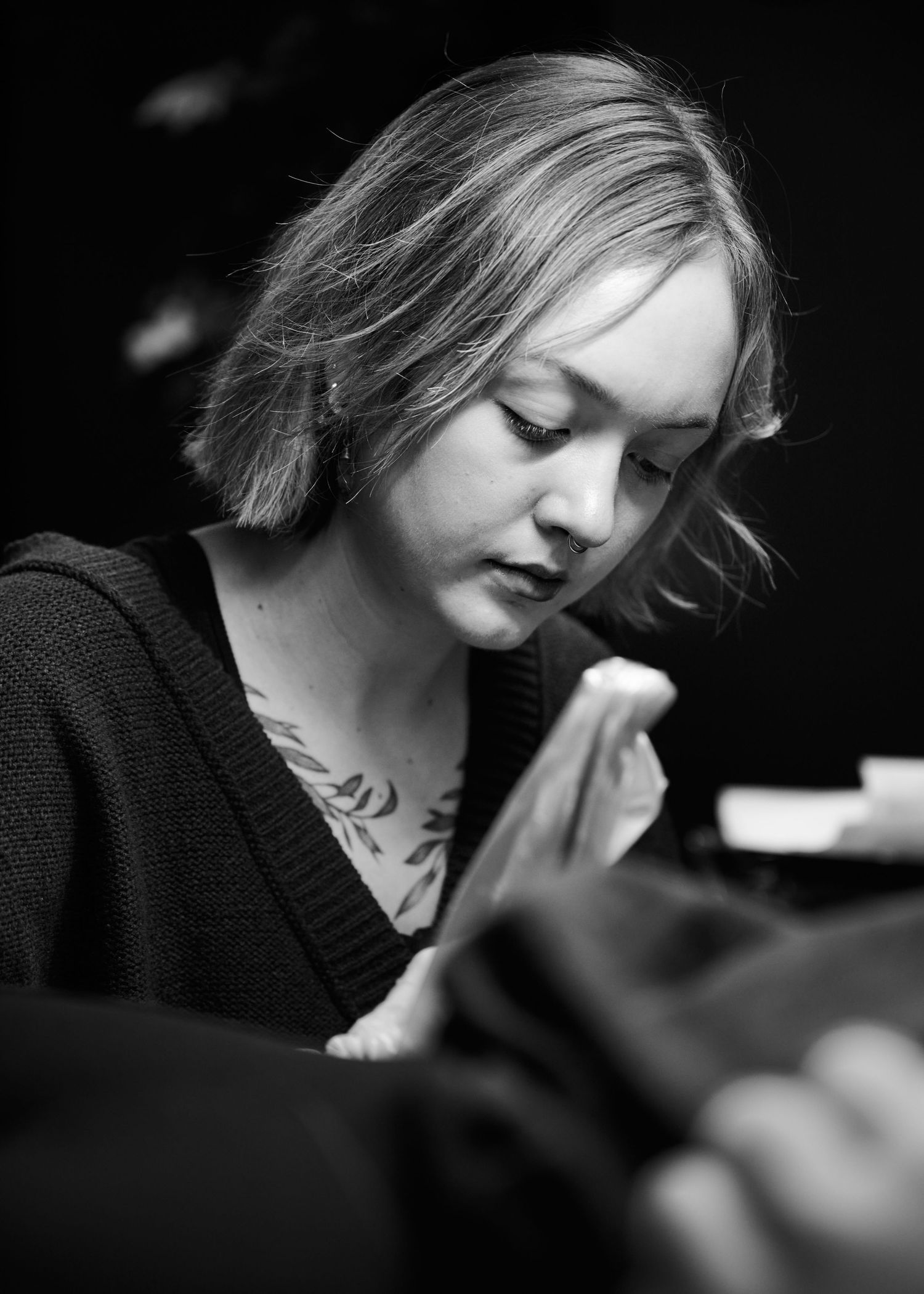 Black and white portrait of someone with closed eyes and neck tattoos wearing a dark cardigan in dim lighting.