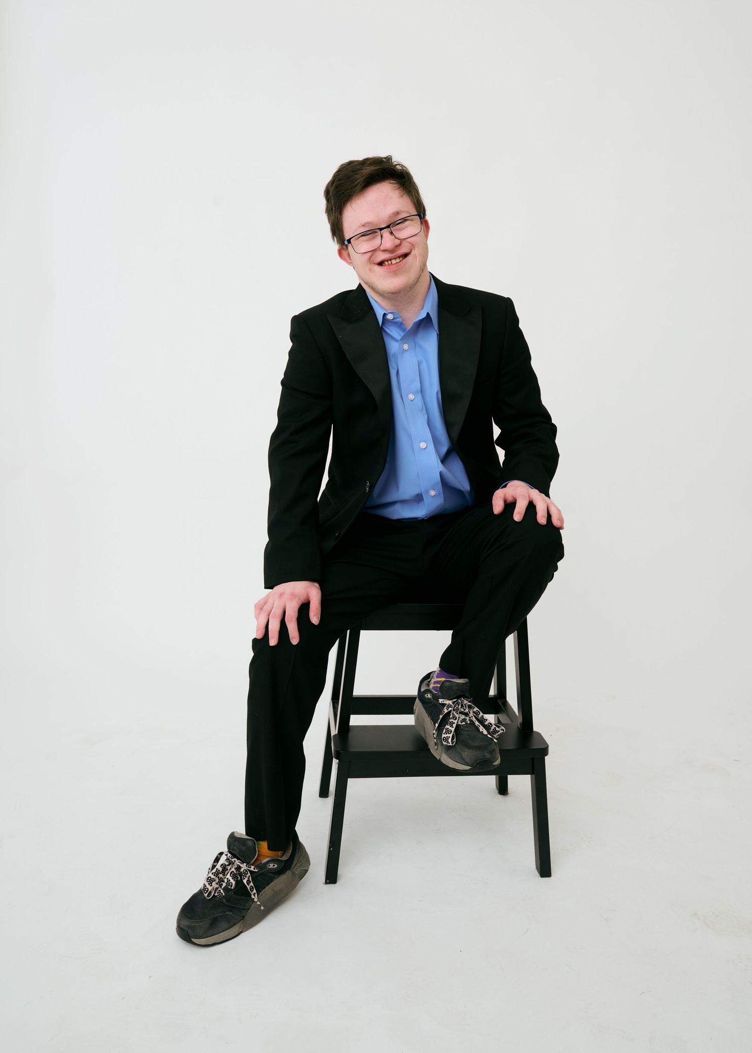 A person in a black suit and blue shirt sits casually in a chair against a white backdrop in four different poses.