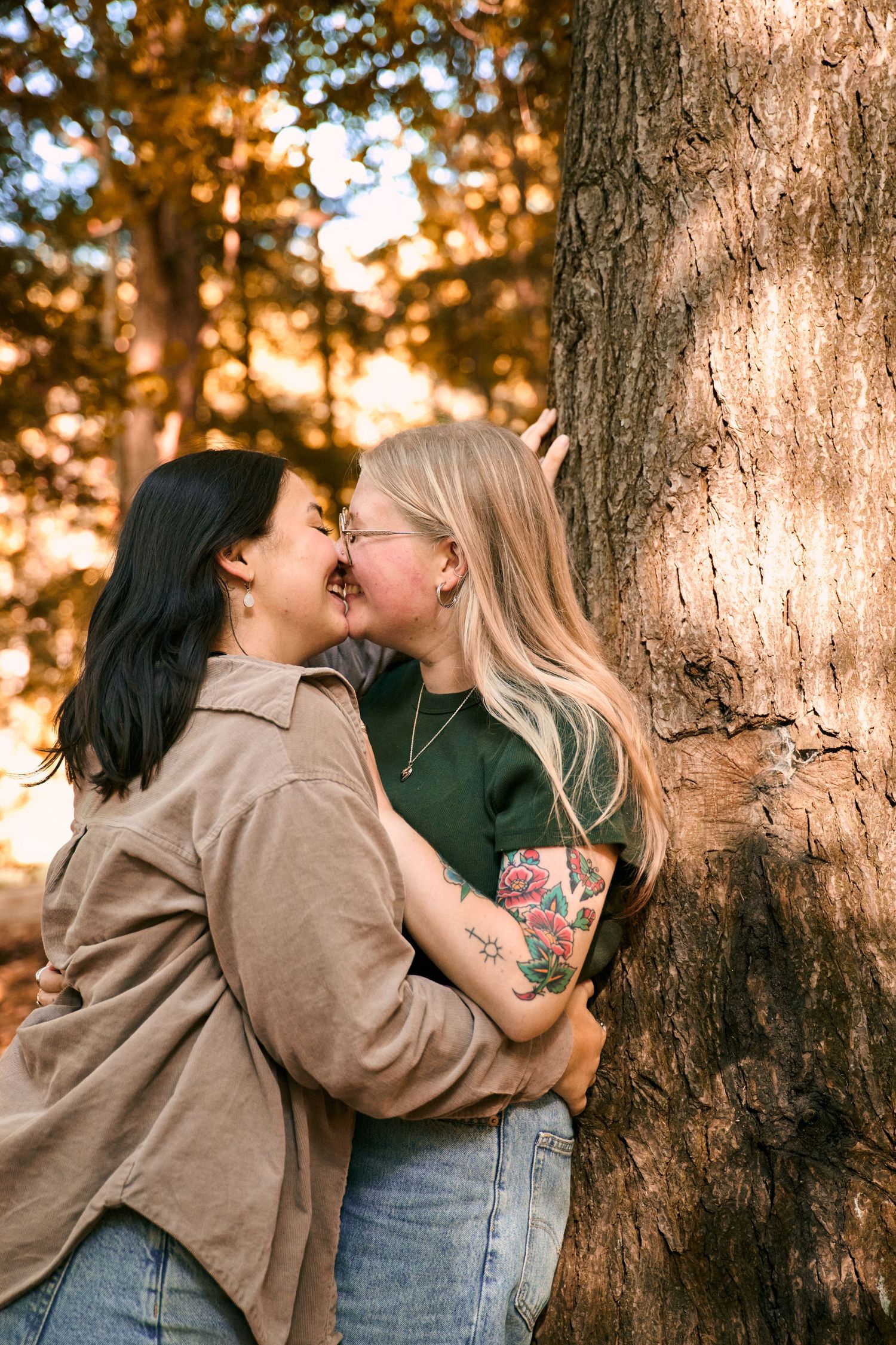 A romantic couple embraces and kisses against a tree trunk during golden hour sunset lighting.