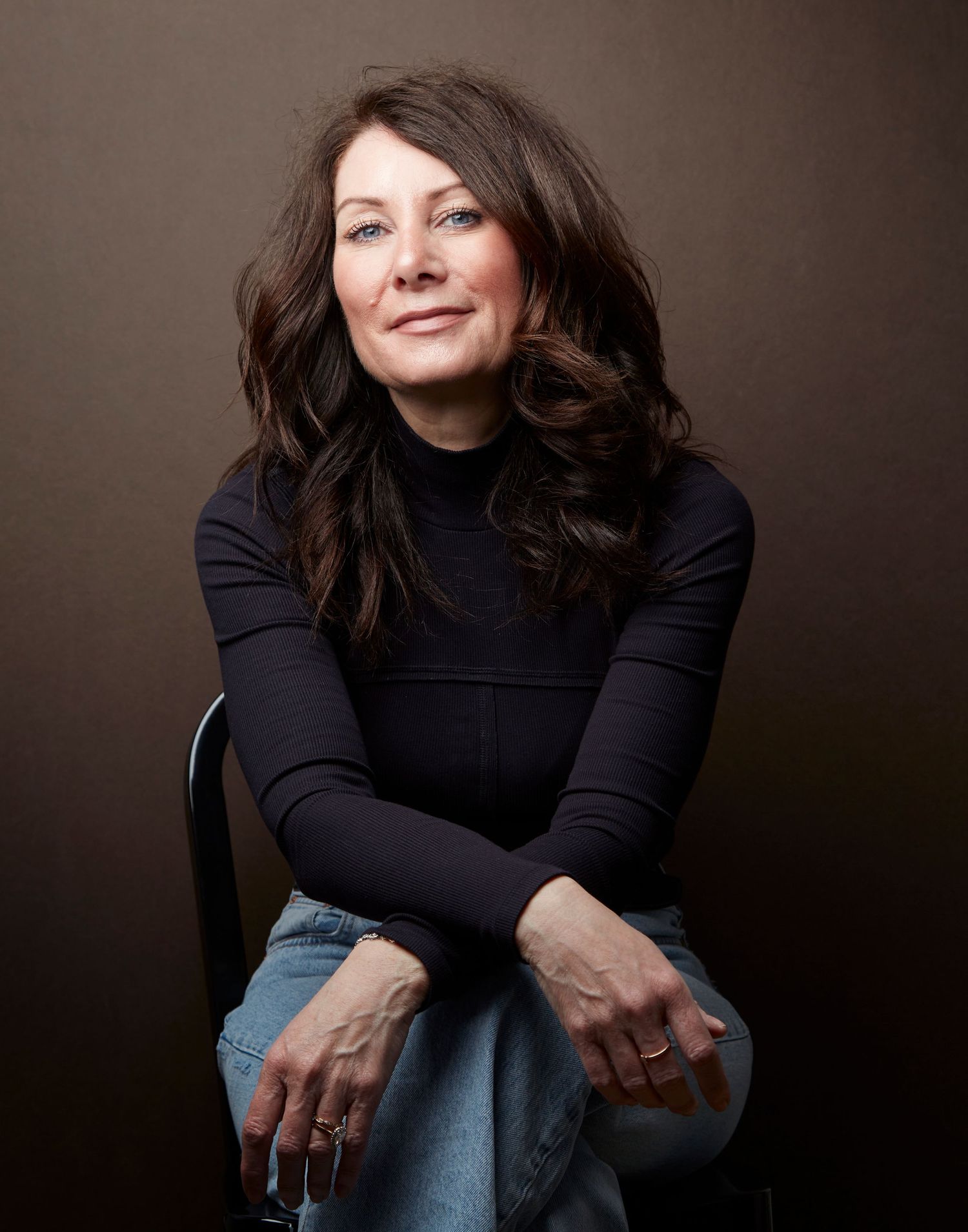A portrait of someone in a black turtleneck sweater and jeans against a dark brown background.