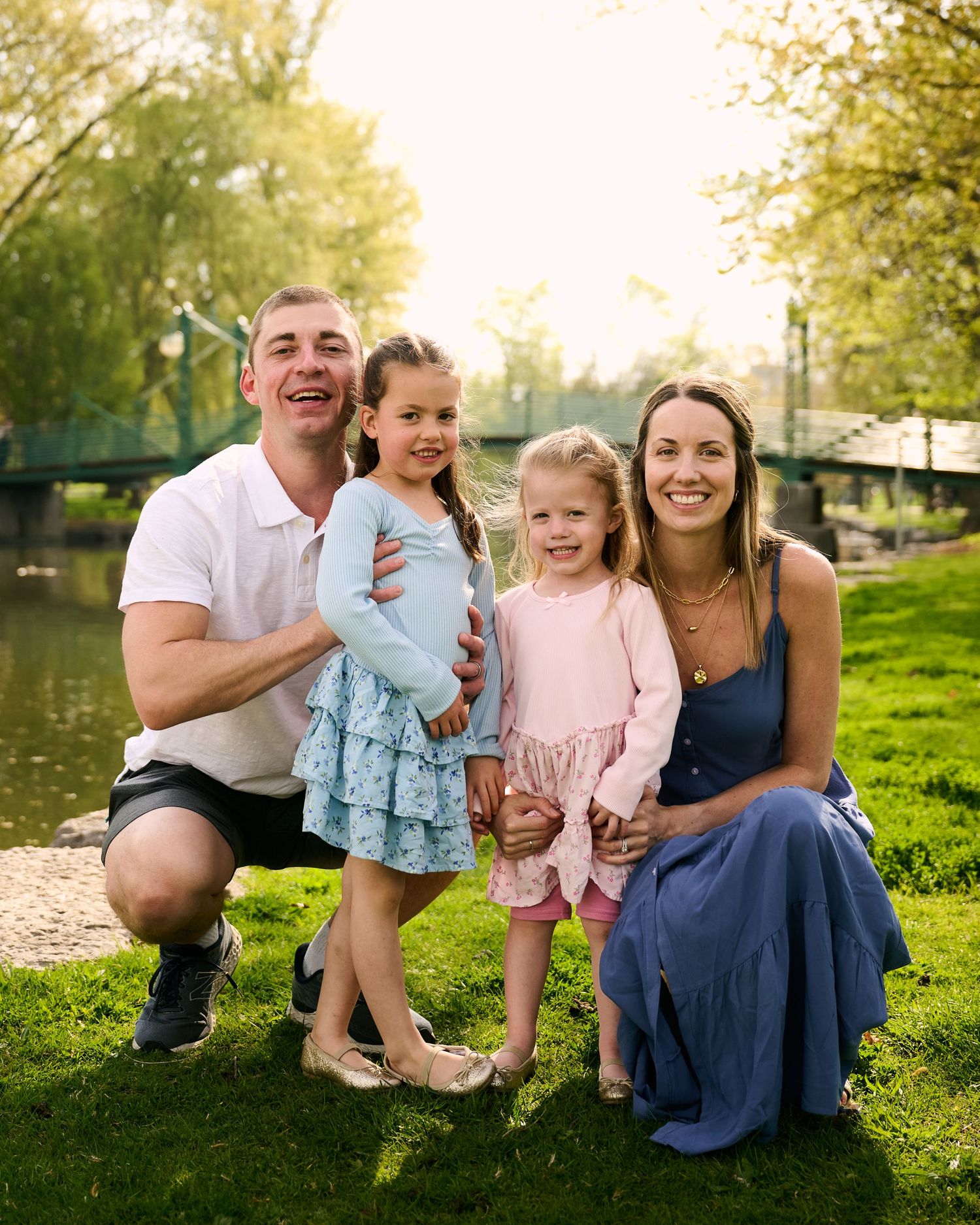 Happy family of four poses outdoors near a pond, parents smiling with two young girls in spring attire.