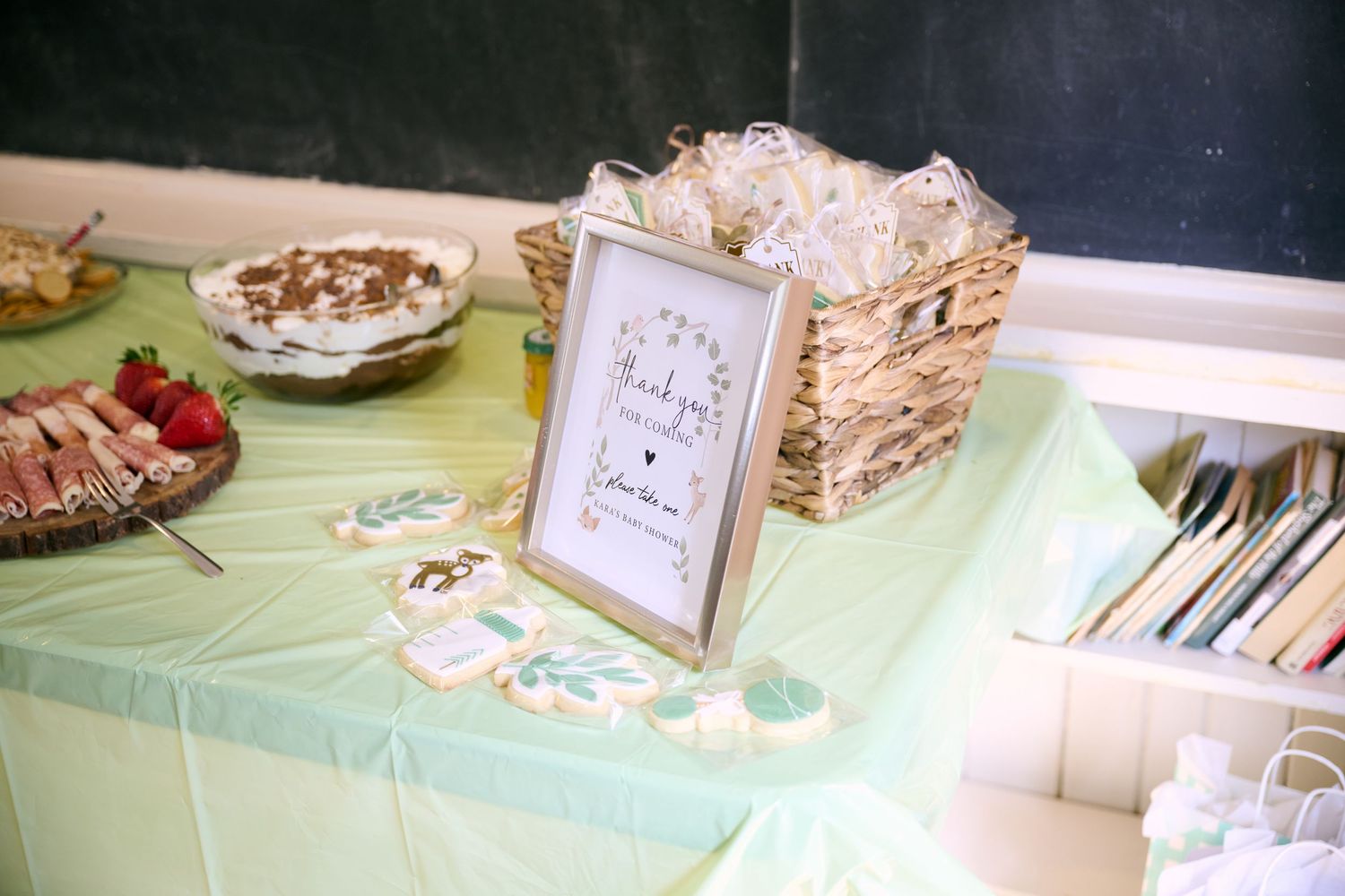 A buffet table with a mint green tablecloth displays a framed sign, woven basket, and party refreshments.