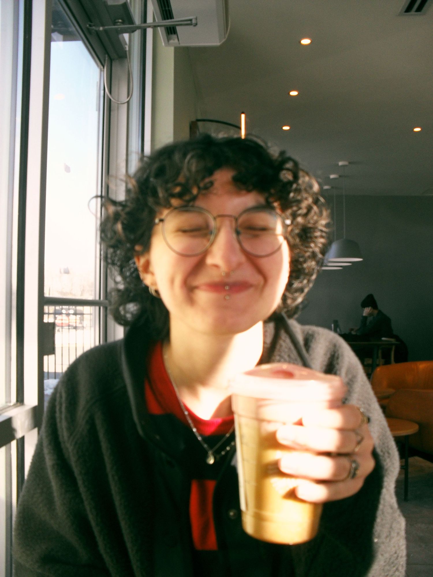 Someone wearing glasses and a dark sweater holds an iced coffee drink while smiling by a window in a cafe.