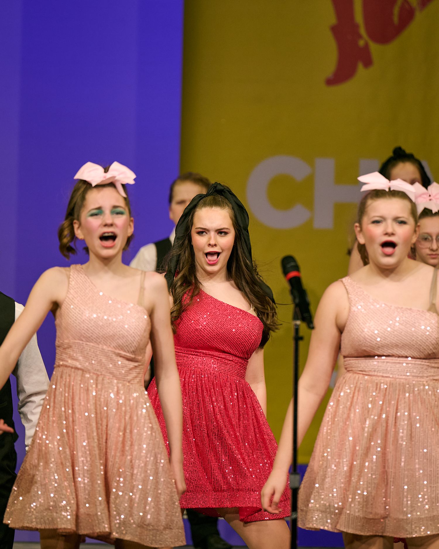 Three girls in pink sequin dresses with bow headbands singing on stage during a performance.