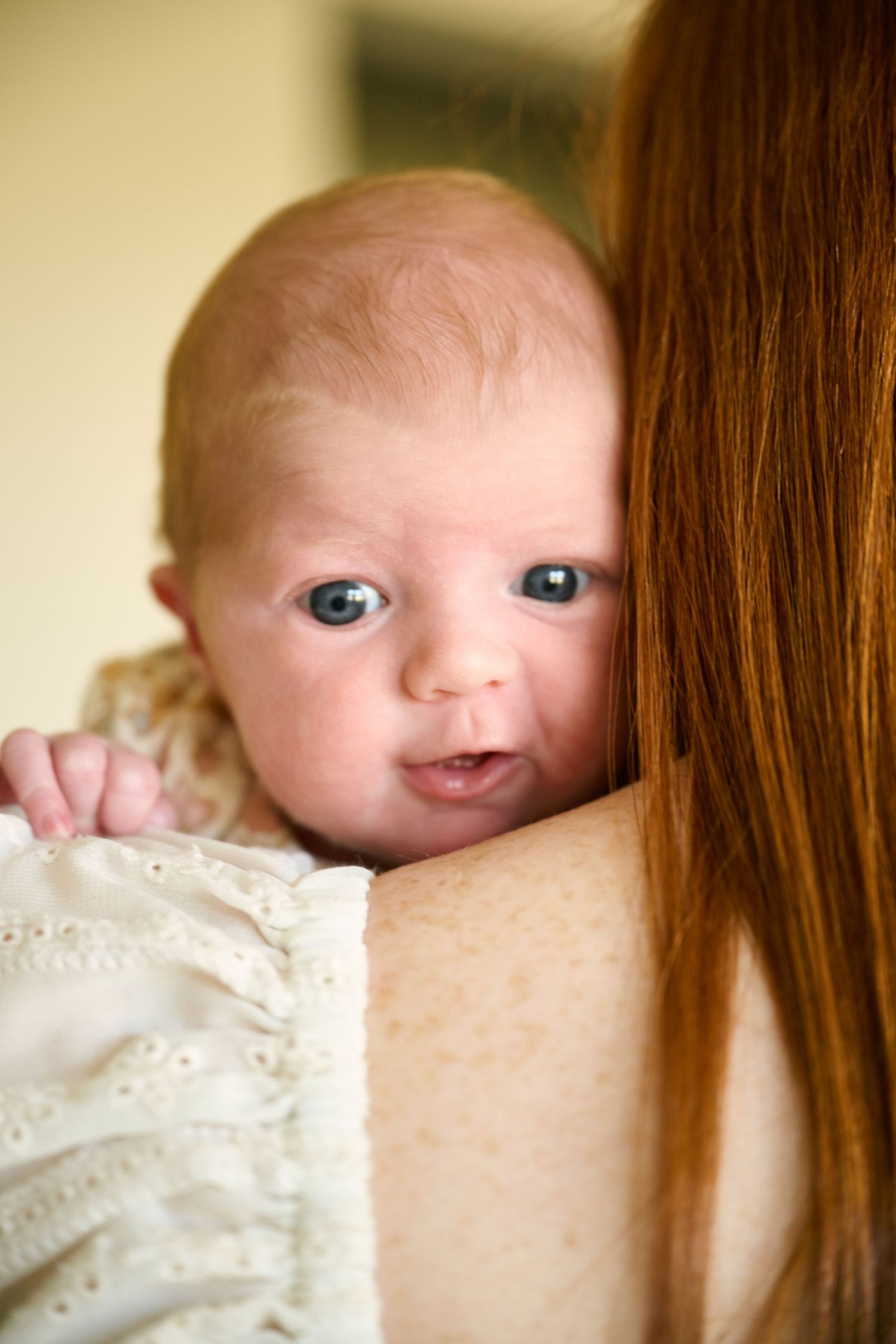 A newborn baby with red hair resting on a redheaded mother's shoulder, wearing a white lace outfit.