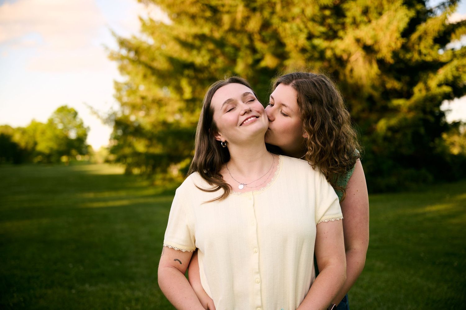 Two young women share a tender moment outdoors, one kissing the other on the cheek in a sunny green park.