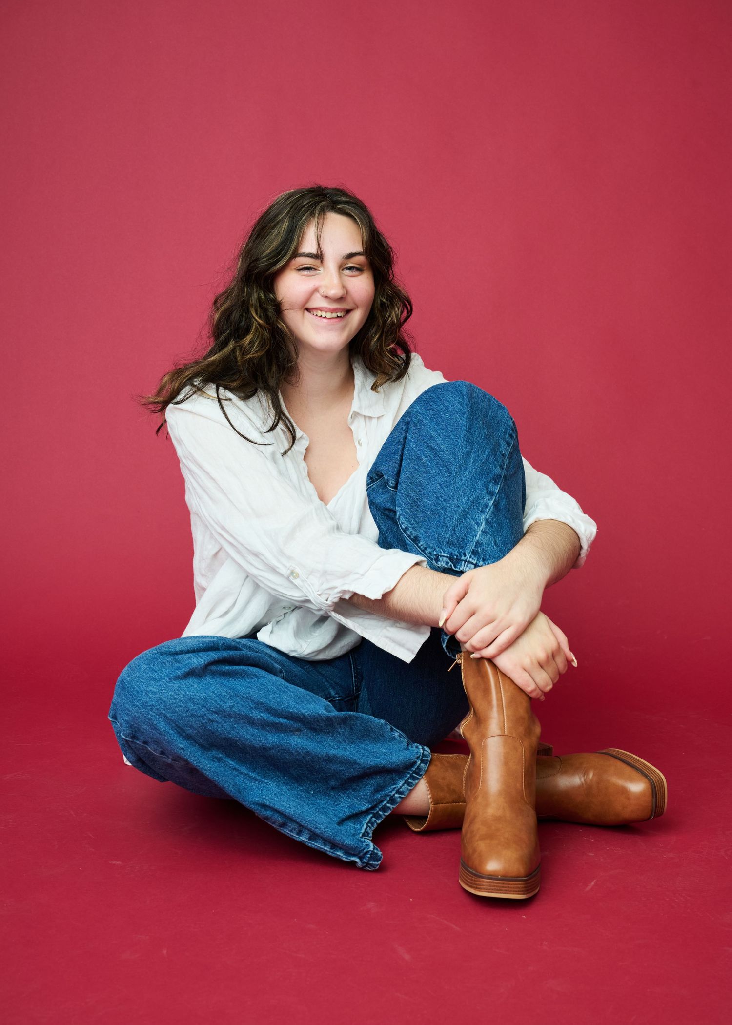 A casual portrait of someone in denim and white shirt sitting cross-legged with brown boots.