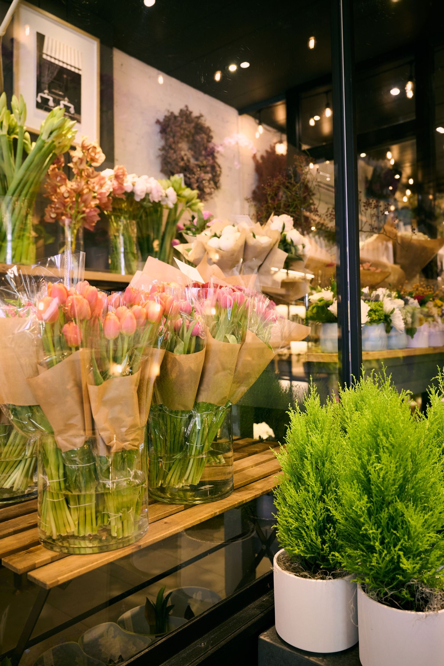 Fresh floral arrangements including pink blooms and potted plants displayed on wooden shelves in an elegant flower shop interior.