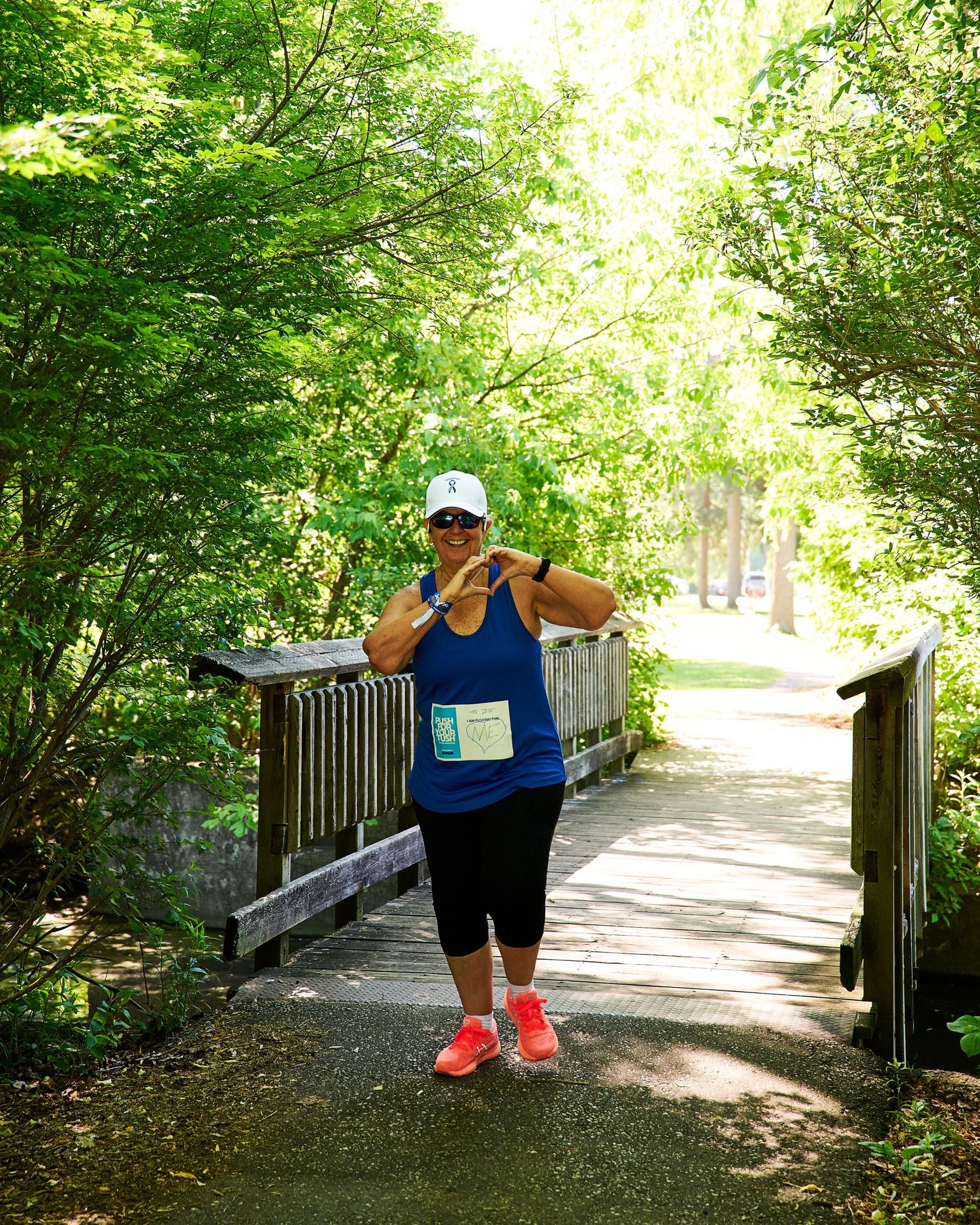 Woman in blue tank top and white cap poses on a scenic tree-lined bridge path wearing bright orange sneakers.