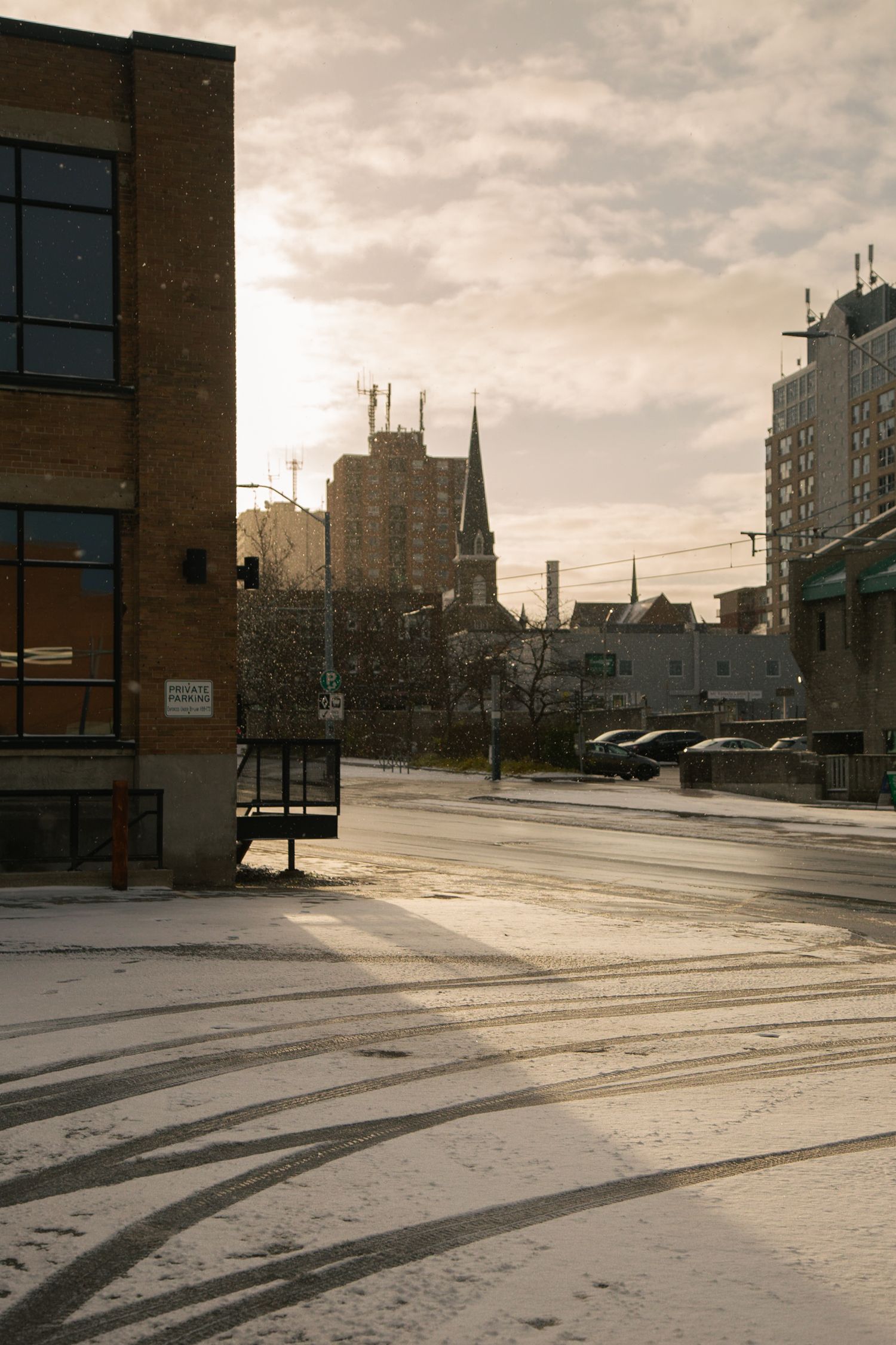 Snow covered urban street at dusk with tall buildings and church spires silhouetted against cloudy sky.