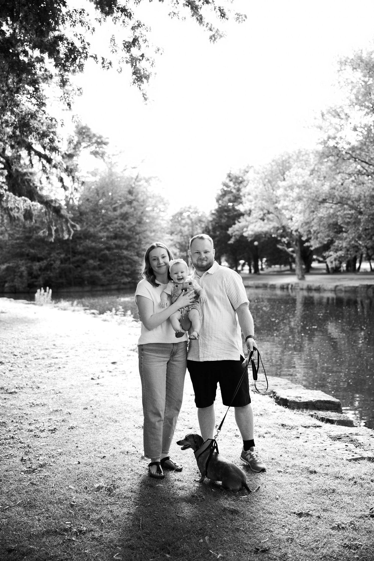 Black and white photo of a smiling family with a baby and dachshund dog posing by a pond in a lush park.