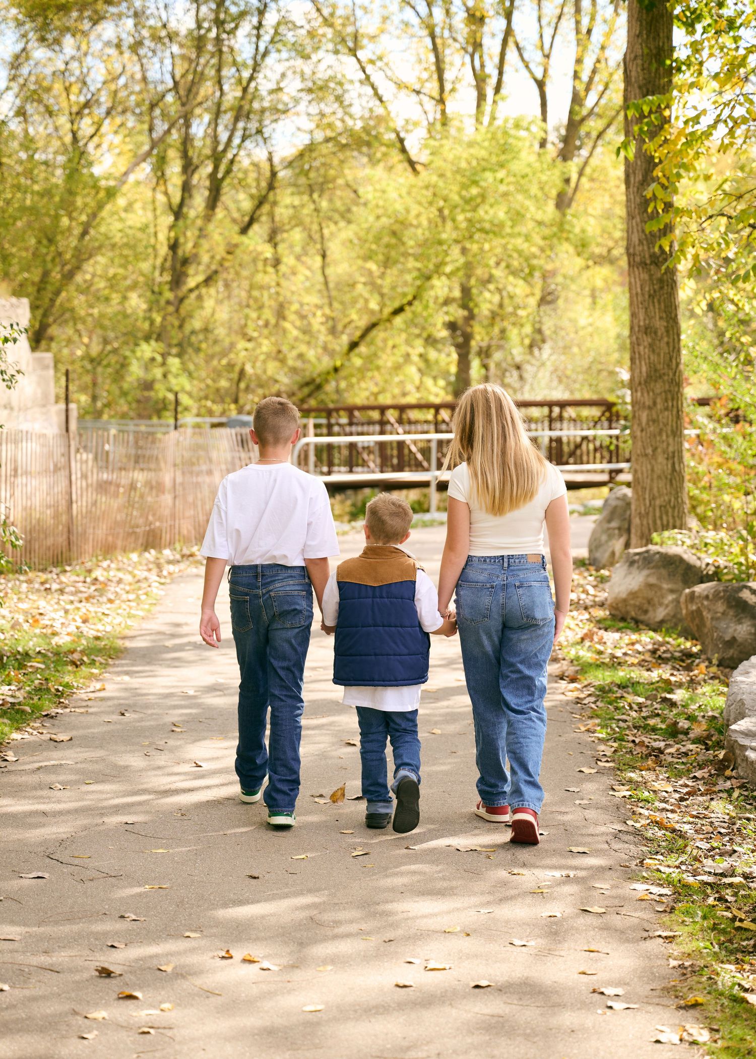 Family walking together on a tree-lined path in autumn with fallen leaves scattered on the ground.