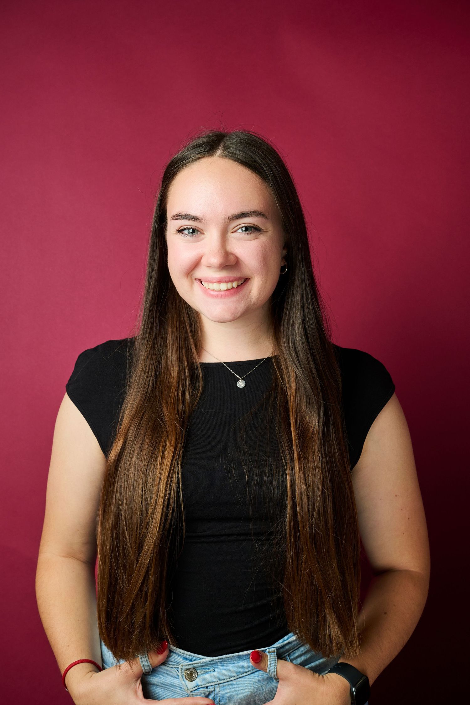 Portrait against burgundy backdrop showing bright smile and long brown hair, wearing black top and blue jeans.