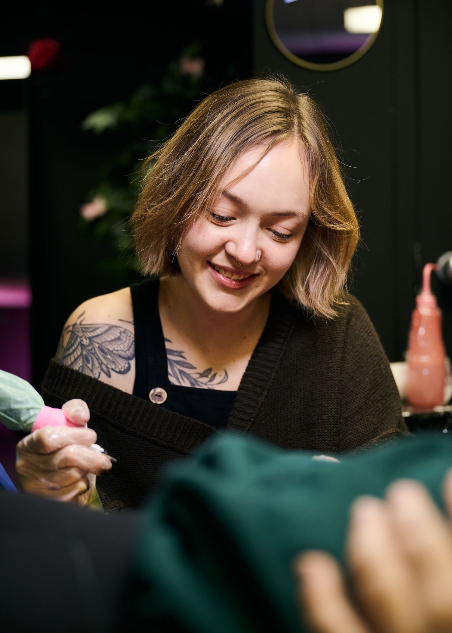 Person with blonde hair and shoulder tattoo smiles while looking down in a black top at a social gathering.