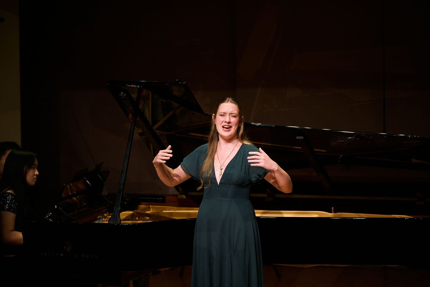 Woman in teal dress performing vocal solo on stage with grand piano and pianist in background.