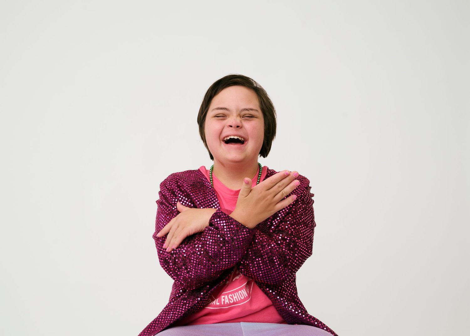 Someone wearing a sparkly purple jacket laughs joyfully while sitting against a white background.