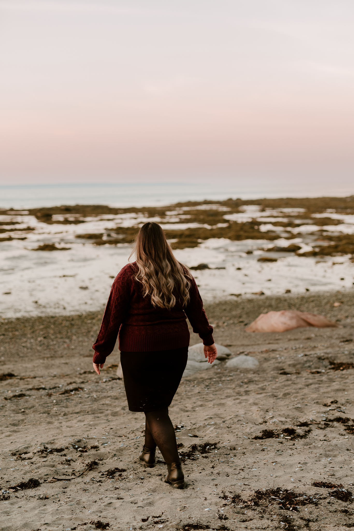 Woman in burgundy top and black skirt walks along a rocky beach at dusk with pastel pink sky.