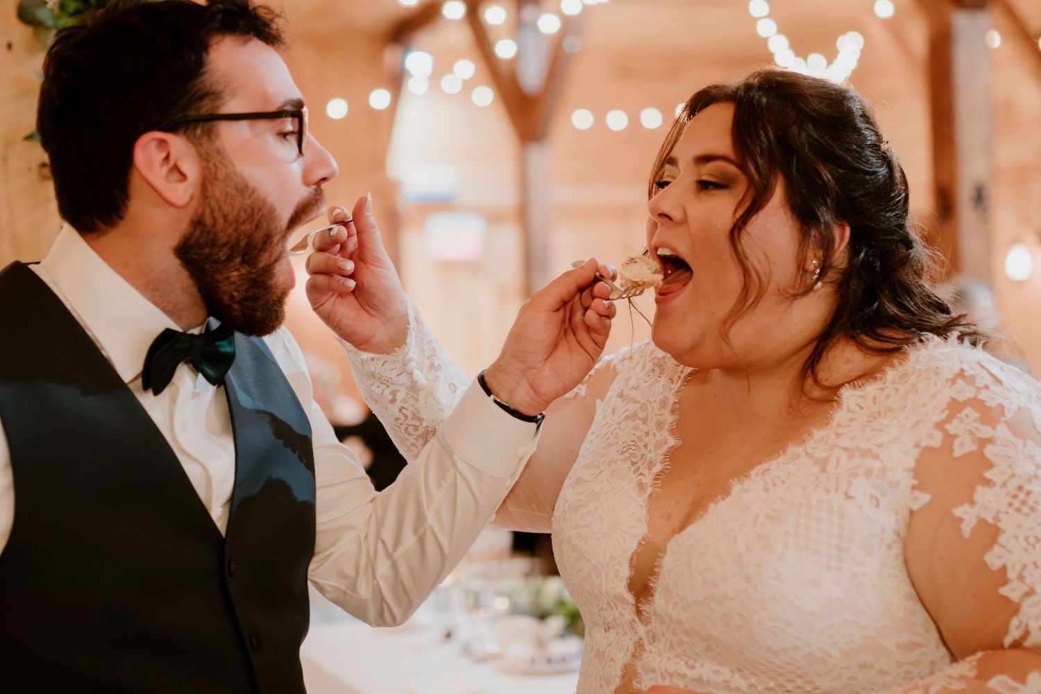 Bride and groom sharing wedding cake during reception in rustic barn venue with warm string lights.