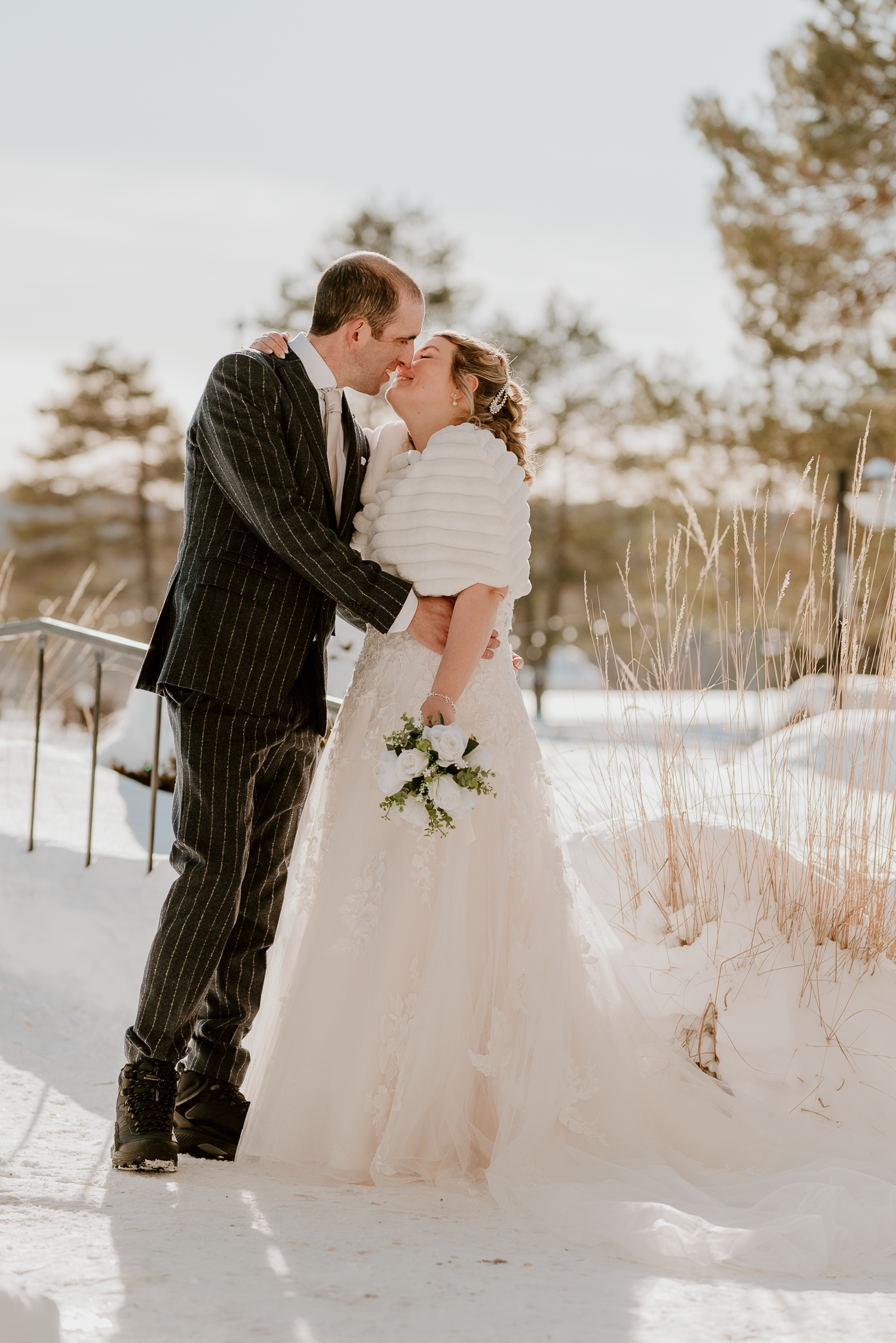 Bride and groom share a kiss in snowy winter landscape, bride in white gown with bouquet, groom in pinstripe suit.