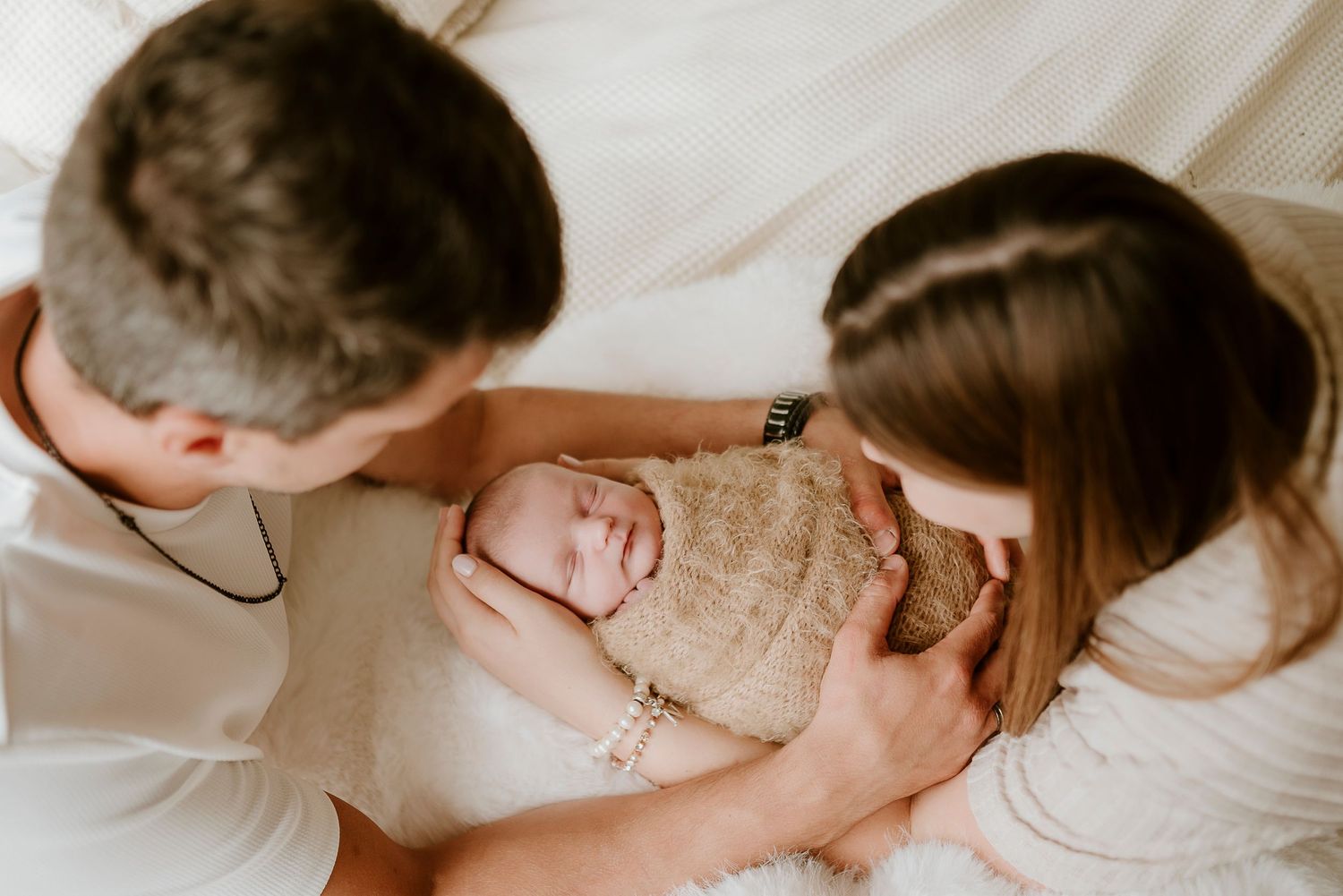 Parents lovingly gaze at their swaddled newborn baby wrapped in a soft beige blanket during a newborn photography session.