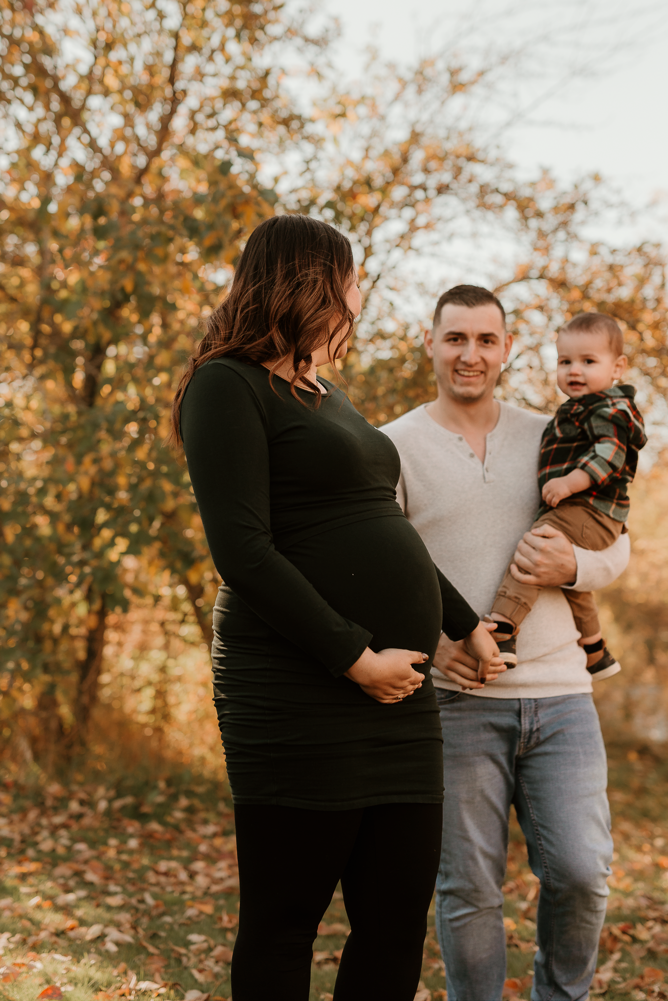 Pregnant woman in black dress poses with smiling man holding toddler during autumn maternity photo session outdoors.